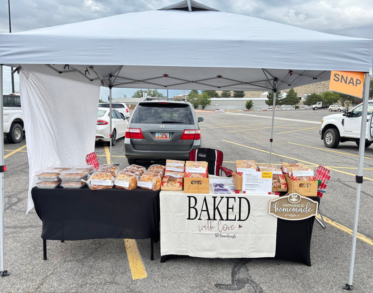 A white pop-up canopy tent set up in Price, Utah Sutherland's parking lot as a  sourdough bread vendor table. Under the canopy, a black tablecloth-covered table displays multiple loaves of bread and packaged baked goods on top of a white table runner that reads 'BAKED with love, SD Sourdough'.